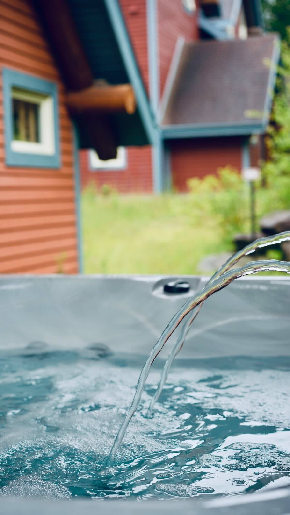 A hot tub with water flowing into it, a wooden house with a blue window in the background.