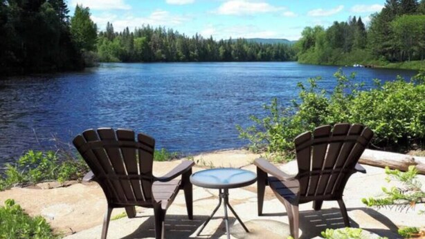 Two Adirondack chairs and a small round table on a stone patio overlooking a lake with a forested shoreline.