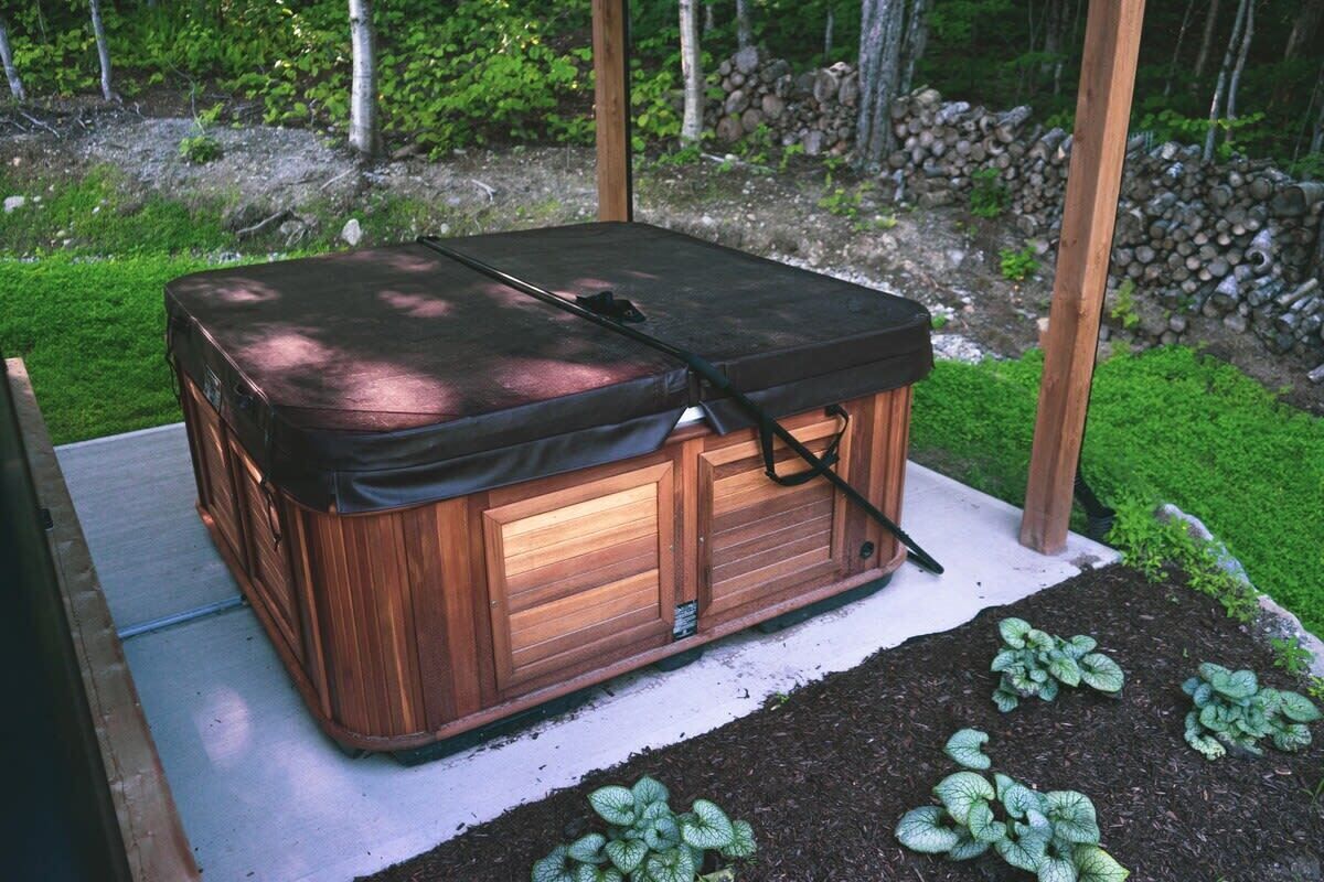 Een hottub met een houten buitenkant en een zwarte afdekking, geplaatst op een betonnen terras omringd door groene planten.