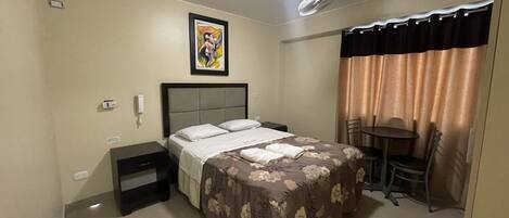Habitación de hotel con cama, escritorio, silla, una mesita, ventilador de techo y un cuadro en la pared.