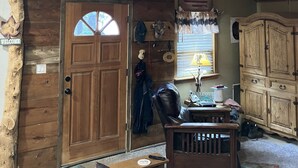 Interior rústico de una cabaña con una puerta de madera, un cartel de bienvenida, un perchero, un sillón de cuero y un armario de madera.