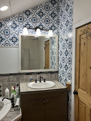 A bathroom with a white sink, a mirror with lights, patterned tiles, and wooden cabinets. - Beautiful Cabin located in the Blue mountain’s, pet friendly and many extras. (Weston)