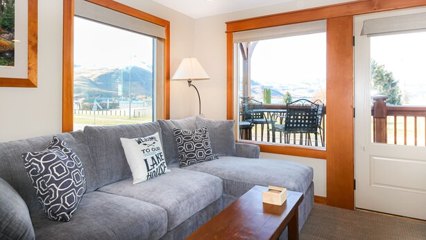 A modern living room with a grey sofa, patterned cushions, a wooden coffee table, and large windows offering a view of outdoor furniture and mountains. - Adrift - Unit 6 (Chelan)