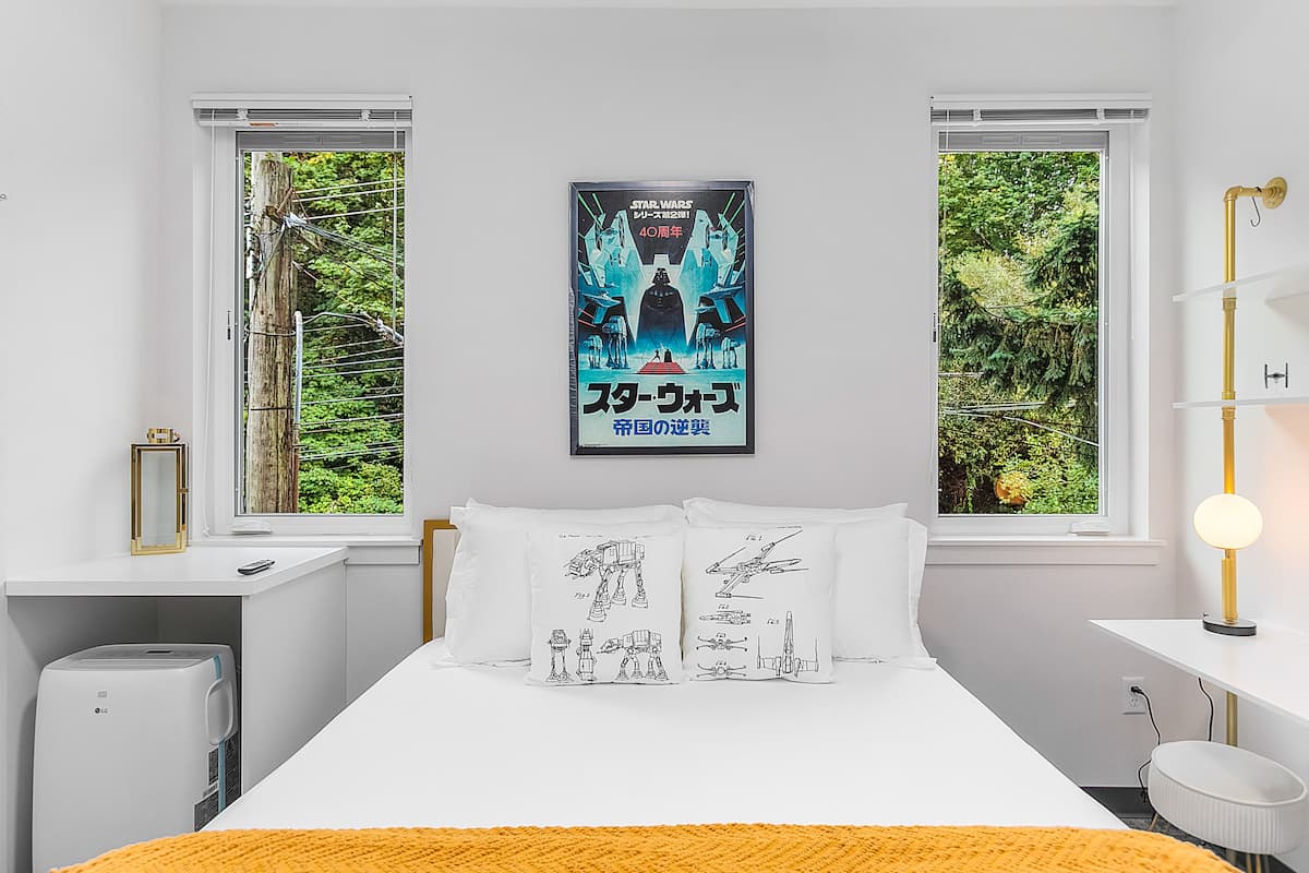 A bedroom with a Star Wars poster, a bed with pillows featuring Star Wars characters, a bedside table, a lamp, and a view of greenery through the windows.
