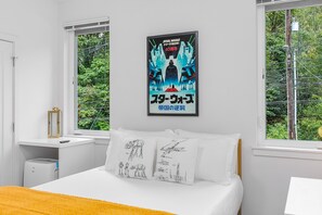 A bedroom with a bed, a bedside table, a nightstand, a poster on the wall, and a window with a view of greenery.