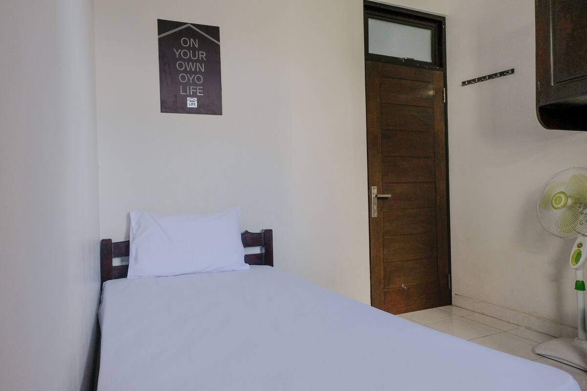 Una cama individual con ropa de cama blanca, cabecera de madera, un letrero en la pared, una puerta de madera y un ventilador de pie.