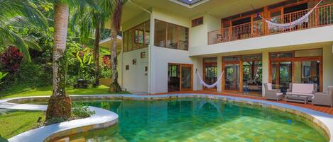 Une maison à deux étages avec une piscine, entourée d’une végétation luxuriante et de palmiers.