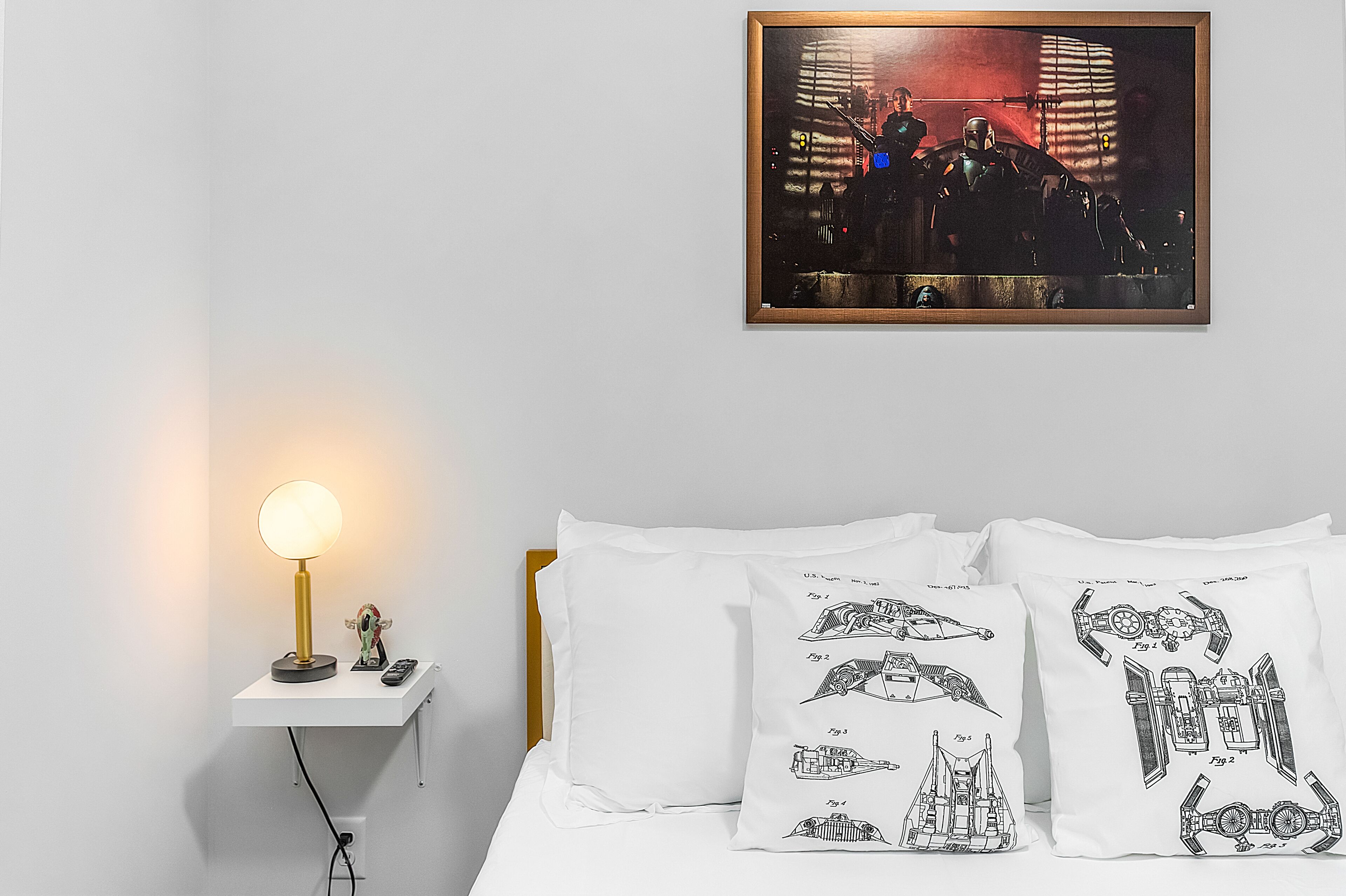 Ein ordentlich eingerichtetes Schlafzimmer mit Nachttischlampe, einem gerahmten Kunstwerk und zwei Kissen mit Raumschiffmotiven.