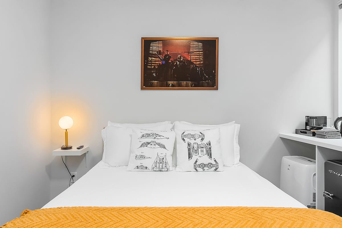 A hotel room with a bed, pillows with space-themed designs, a nightstand with a lamp, a framed picture on the wall, and a small shelf with a coffee maker and other items.