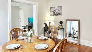 Uma sala de jantar com uma mesa redonda posta para quatro pessoas, cadeiras de madeira, um vaso com flores e um quadro na parede.