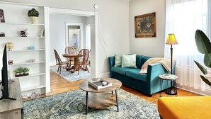 Uma sala de estar com sofá azul, mesa de centro, estante de livros, área de jantar com cadeiras e uma luminária.