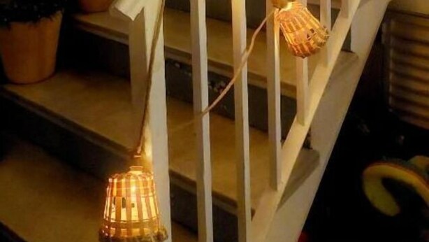 Eine Treppe mit Korblampen, die an der Geländer befestigt sind, Blumentöpfe auf der Zwischenebene und ein Fenster mit Jalousien im Hintergrund.