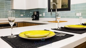A modern kitchen with a white countertop, two yellow plates, and two wine glasses. - Wariruri 1-14 BRAND NEW Apartment, private patio! (Noord)