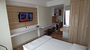 Una habitación de hotel moderna con televisión de pantalla plana montada en un panel de madera, una cama con ropa blanca, un pequeño espacio con cocina que incluye un lavamanos y armarios, y un armario de madera.