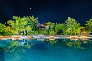 A nighttime pool area with clear water, palm trees, and a building in the background.