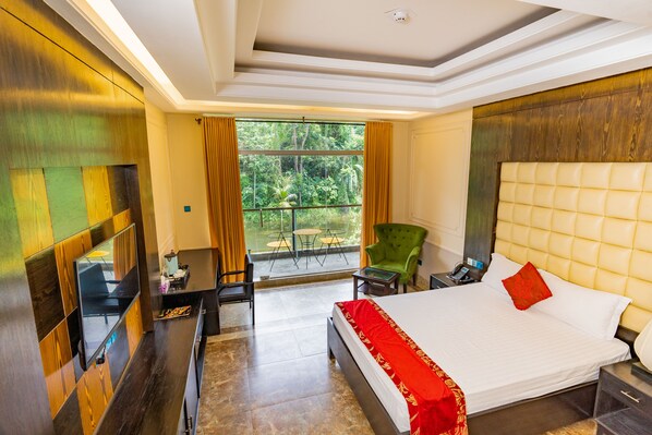 A hotel room with a large bed, a green armchair, a small table, and a balcony with a view of greenery.