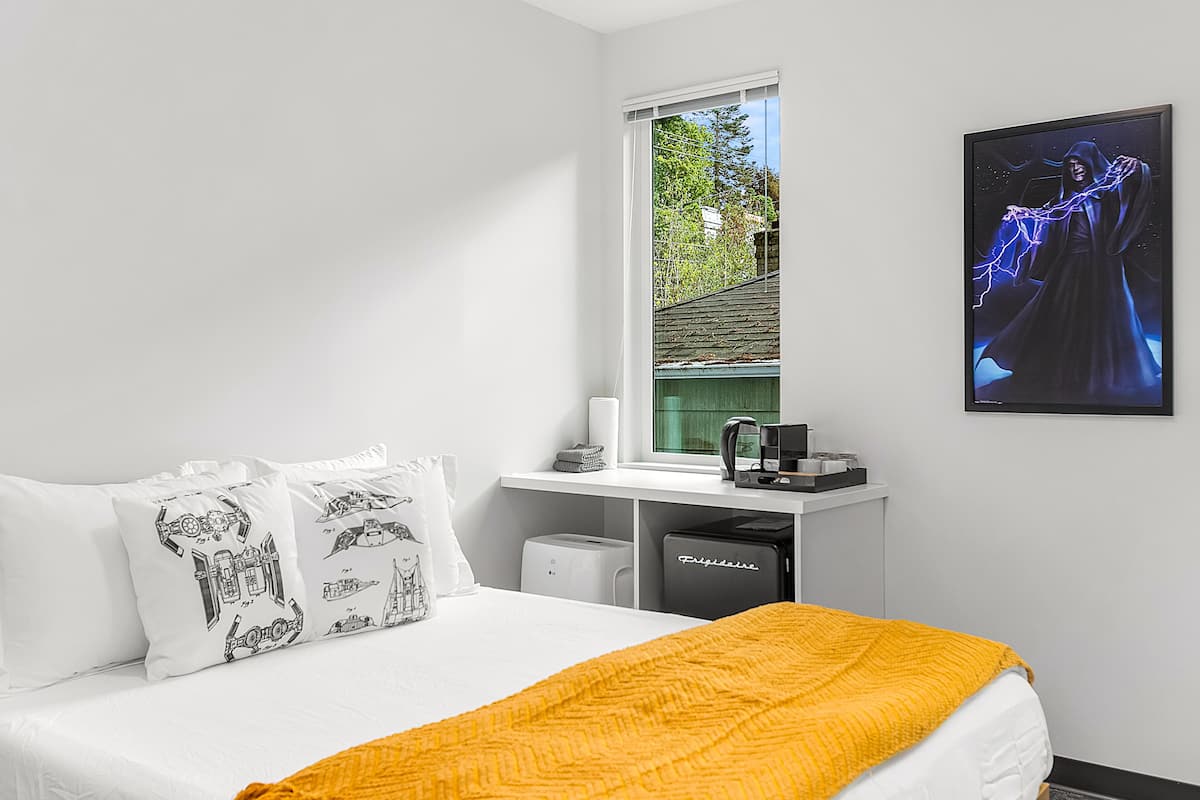 A bedroom with a bed, white pillows, a yellow blanket, a window, a desk with a computer, and a framed picture on the wall.