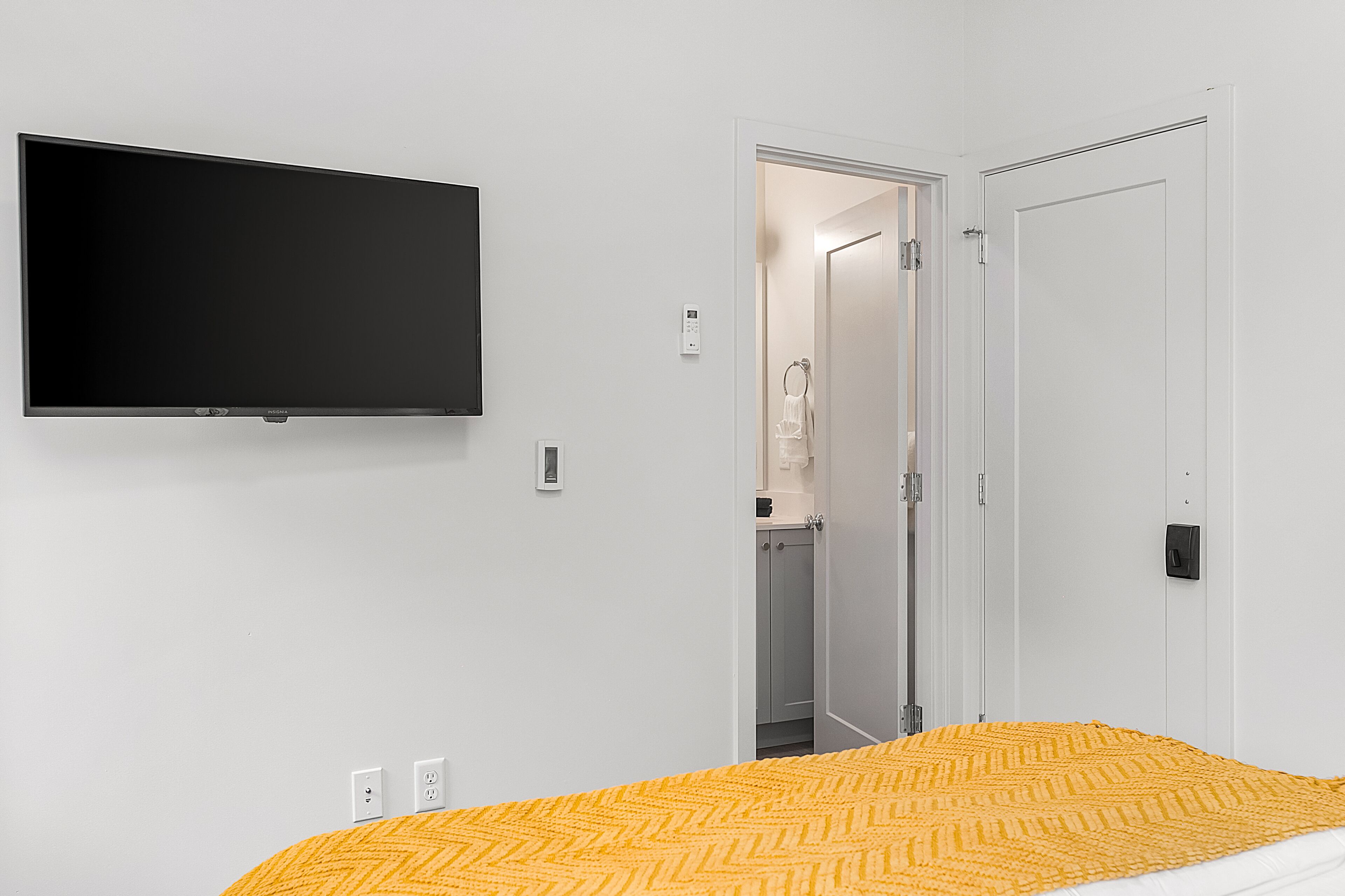A bedroom with a bed, a television mounted on the wall, a door leading to a bathroom, and a power outlet on the wall.