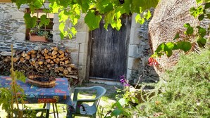 A rustic outdoor seating area with a table covered by a patterned tablecloth, a basket of blueberries, and a stack of firewood against a stone wall. - La Bergerie: Gîte Familial et Chaleureux Dans le Parc National des Cévennes (MALONS-ET-ELZE)
