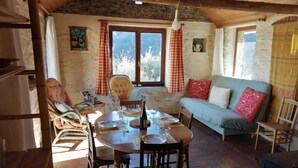 A rustic dining area with a wooden table set for two, surrounded by chairs. A sofa with patterned cushions is against the wall, and a window with curtains is visible. - La Bergerie: Gîte Familial et Chaleureux Dans le Parc National des Cévennes (MALONS-ET-ELZE)