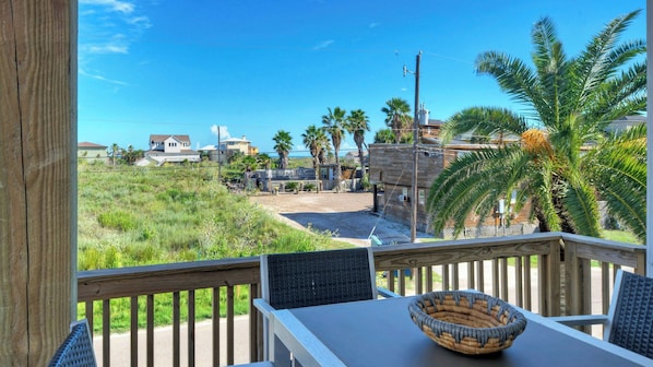 A wooden deck with a table and chairs, overlooking a grassy area with palm trees and houses.