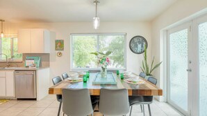 A modern kitchen with a dining area, featuring a wooden table, chairs, a clock, and a window with blinds. - Sea Glass Secrets by Avantstay 3 Mins to Beach (Key West)