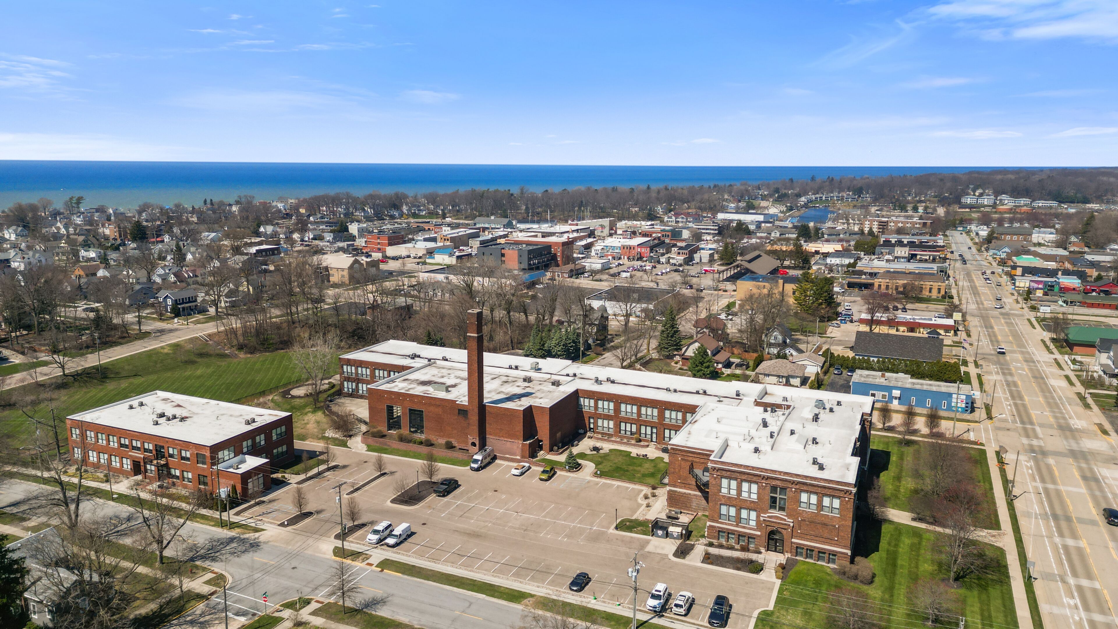 An aerial view of a town with a prominent industrial building featuring a tall chimney, surrounded by residential areas and a body of water in the background.