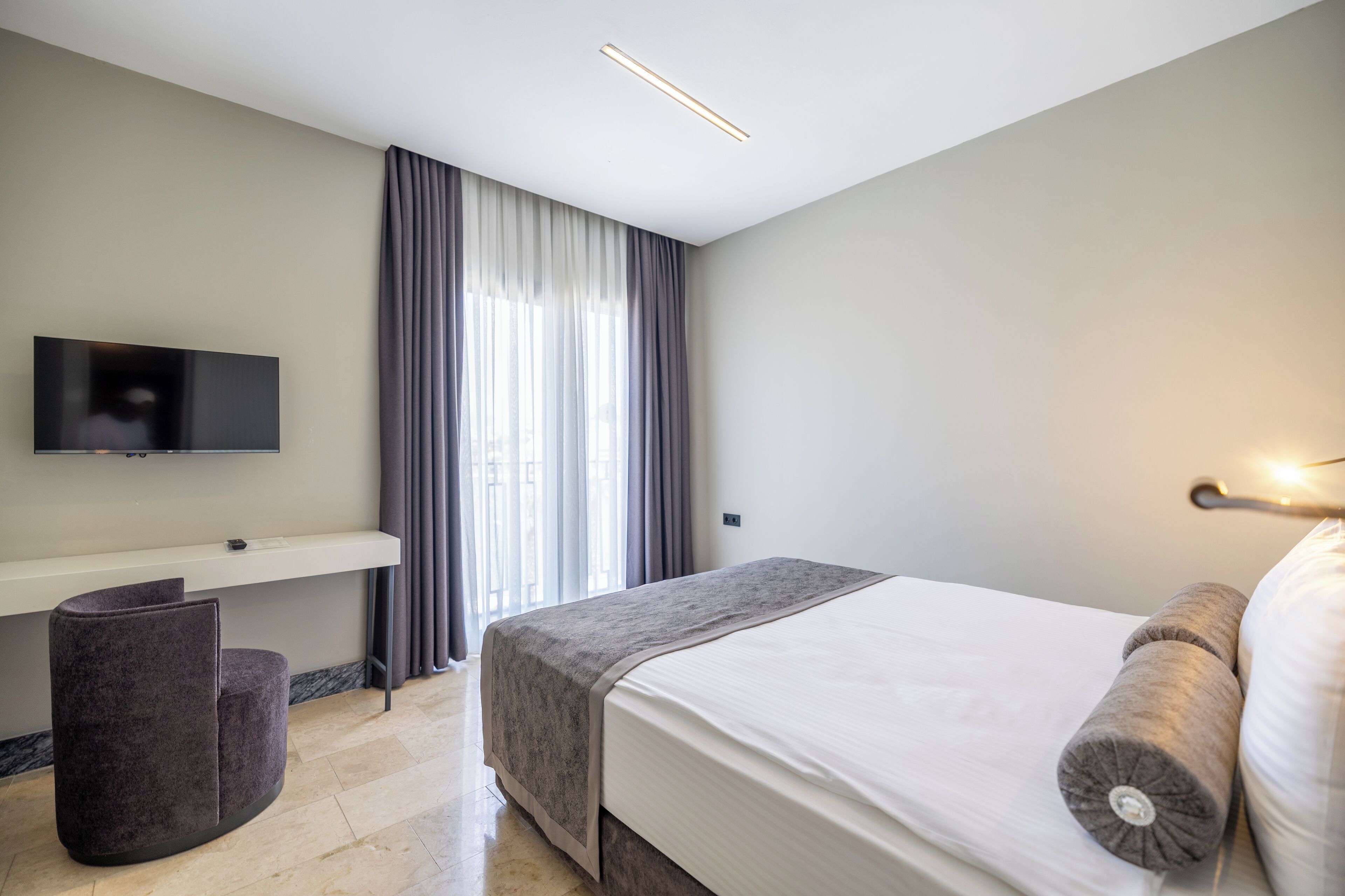 A modern hotel room with a bed, a flat-screen TV, a small seating area, and a window with curtains.