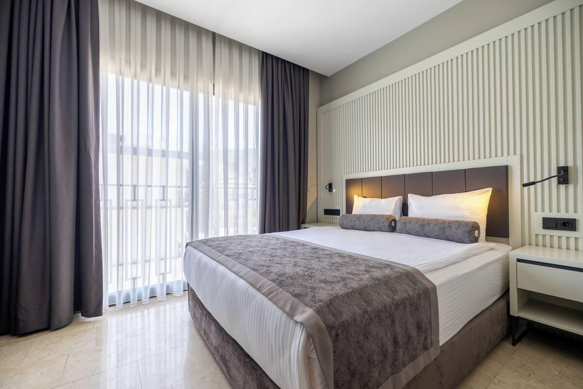 Moderni hotellihuone, jossa on suuri sänky, yöpöytä ja näkymä kaupunkiin ohuiden verhojen läpi.