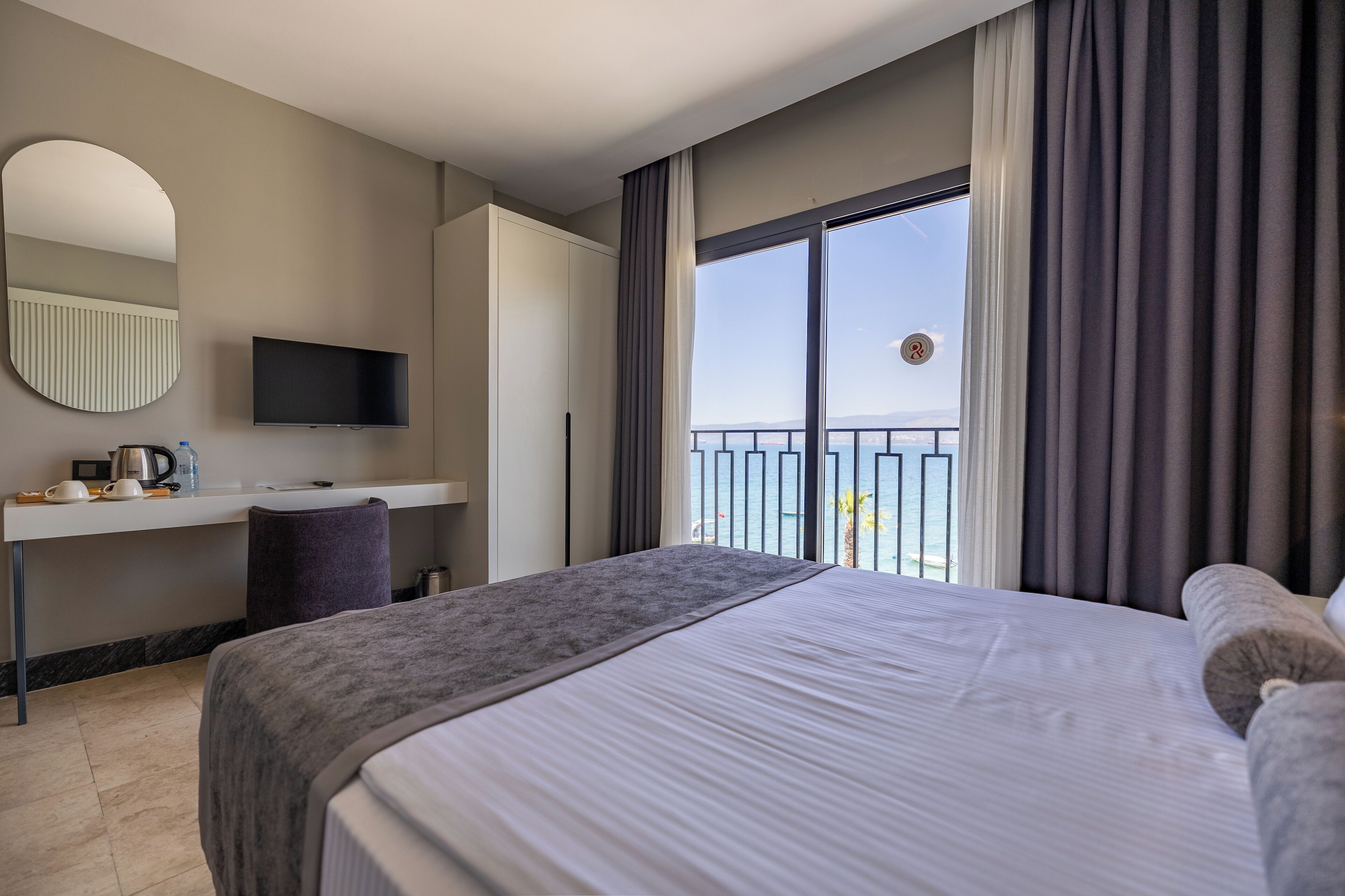 A hotel room with a large bed, a desk with a flat-screen TV, a view of the sea through a balcony, and a round mirror on the wall.