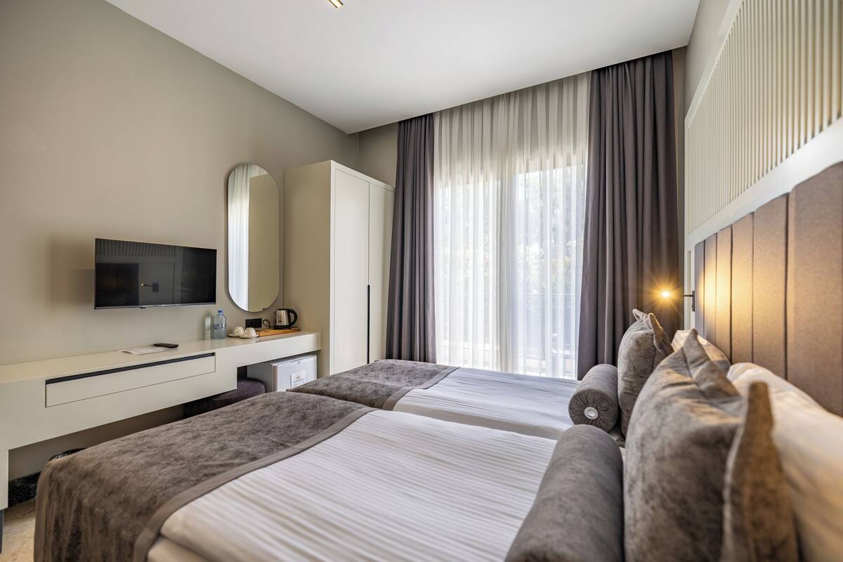 Hotellihuone, jossa on kaksi sänkyä, televisio, työpöytä ja suuri ikkuna, jossa on verhot.