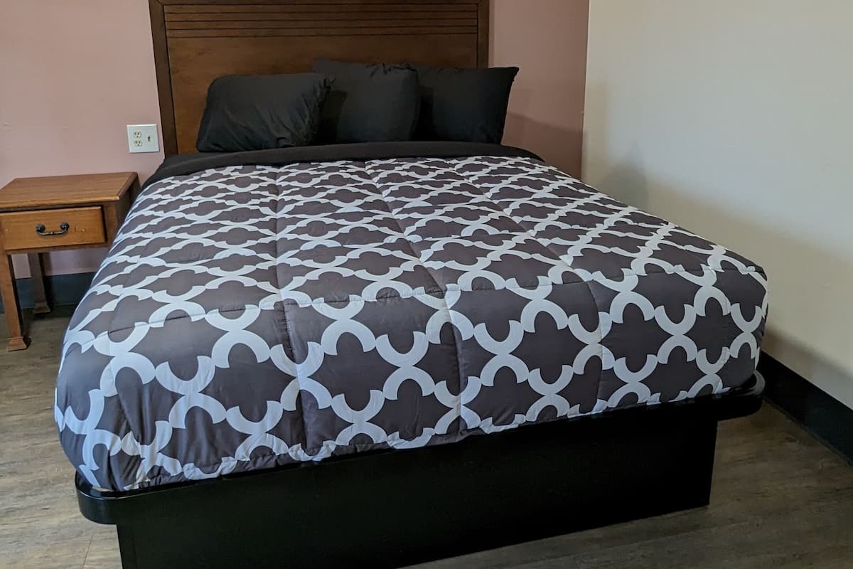Un lit simple avec une couvre-lit à motifs, une tête de lit en bois, une table de chevet avec un tiroir et un luminaire fixé au mur.