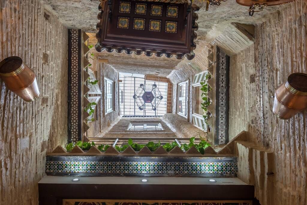 Um corredor estreito e ricamente decorado, com paredes estampadas e chão de azulejos, que leva a uma área central com um desenho geométrico no teto.