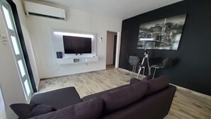A modern living room with a dark-colored sofa, a flat-screen TV mounted on a white entertainment unit, and a cityscape painting on the wall. - Casa Bird Cage in Fajardo's Paradise, a rainforest like retreat! (fajardo)