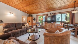 Un salon chaleureux avec un plafond en bois, un grand téléviseur à écran plat et un coin repas comprenant une table et des chaises.