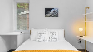A bedroom with a white bed, a window with blinds, a bedside table with a lamp, and a wall with a framed picture. - Jabba's Palace (Seattle)