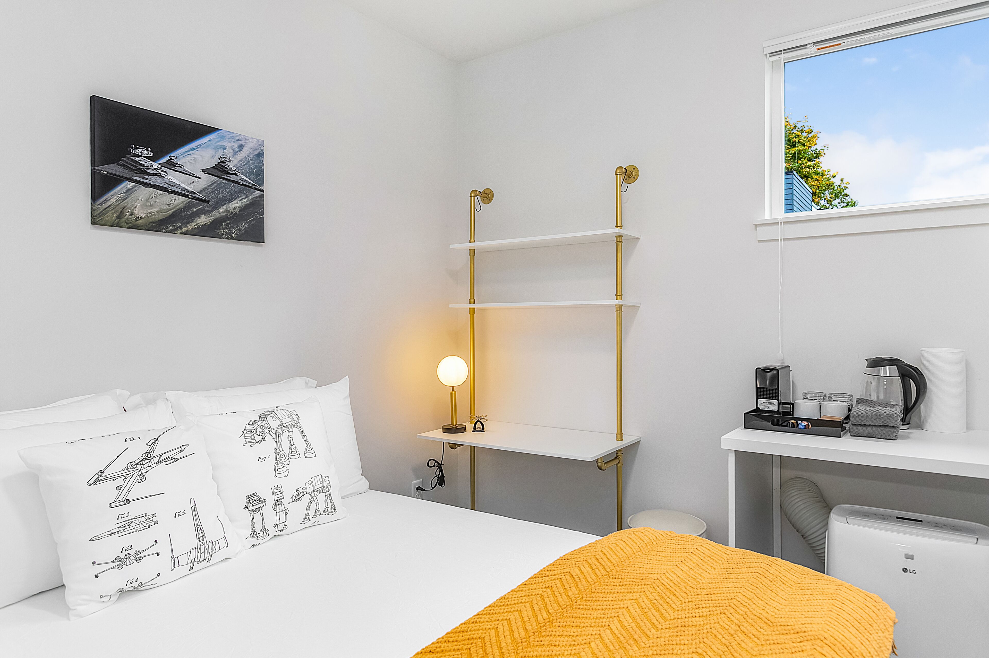 A hotel room with a bed, a bedside table with a lamp, a shelf unit, and a window with a view of the sky.