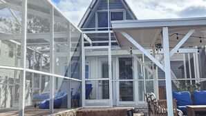 Un espace extérieur abrité, doté d’une structure en verre, avec une maison bleue en arrière-plan et du mobilier d’extérieur.