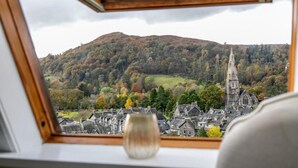 A view from a window with a wooden frame, overlooking a town with a prominent church spire and a hill with autumn foliage.