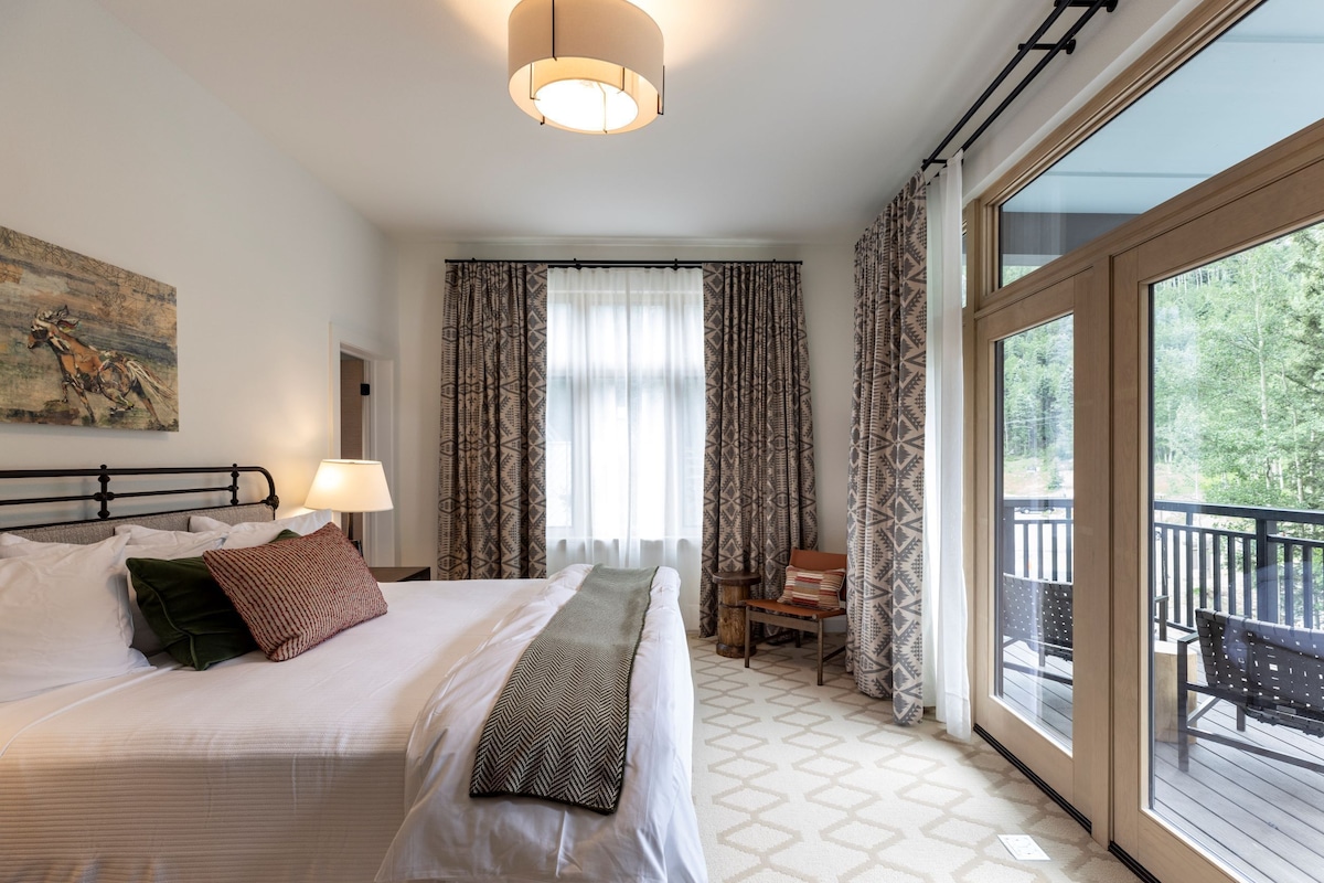 A bedroom with a large bed, a bedside table with a lamp, a window with patterned curtains, and a view of the outdoors.