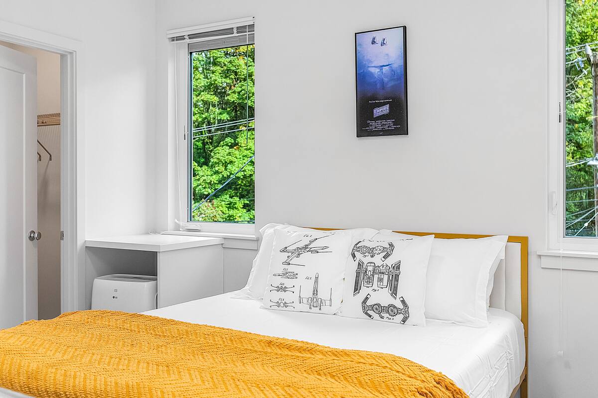 A bedroom with a bed, a window with a view of greenery, a white nightstand, and a wall-mounted poster.