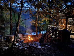 A campfire surrounded by chairs and debris, with a forested backdrop and a cabin in the distance. - Lakefront cabin with dock, hot tub, fire pit!! (Plymouth)