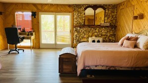Ein Schlafzimmer mit einem Bett, einem Schreibtisch und einem Stuhl. Eine Steinwand und eine Holzbalkendecke sind zu sehen.