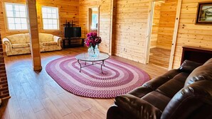 Ein gemütliches Interieur einer Holzblockhütte mit einem Wohnzimmer, das eine braune Ledersofa, einen Glastisch und einen rosa Teppich sowie einen Fernseher auf einem Holzständer zeigt.