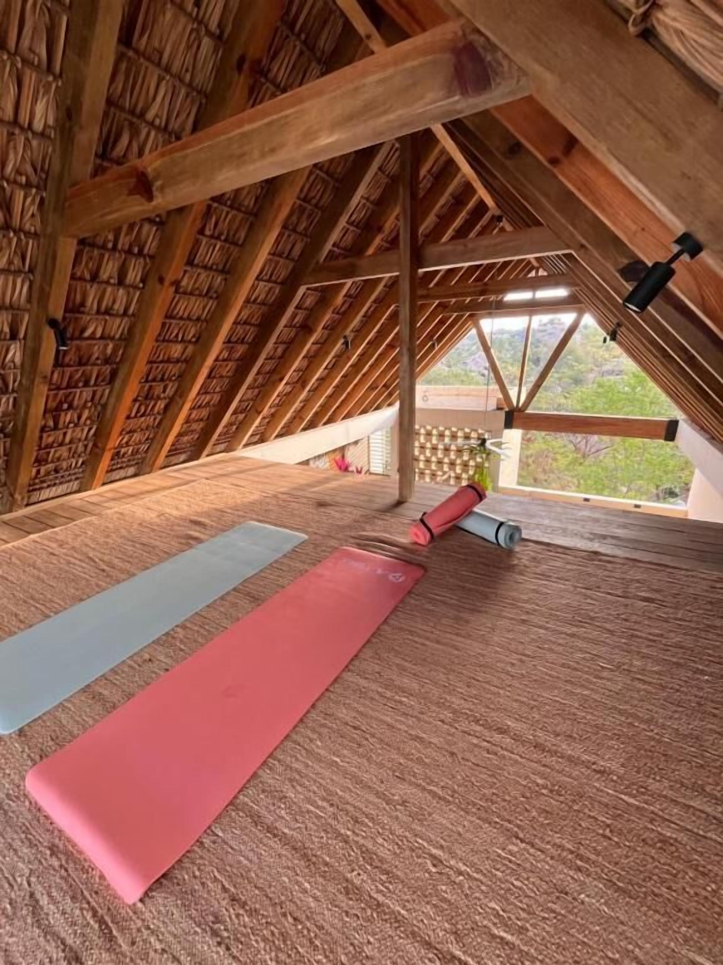 An attic room with a sloped wooden ceiling, a large window, and two yoga mats on the floor.