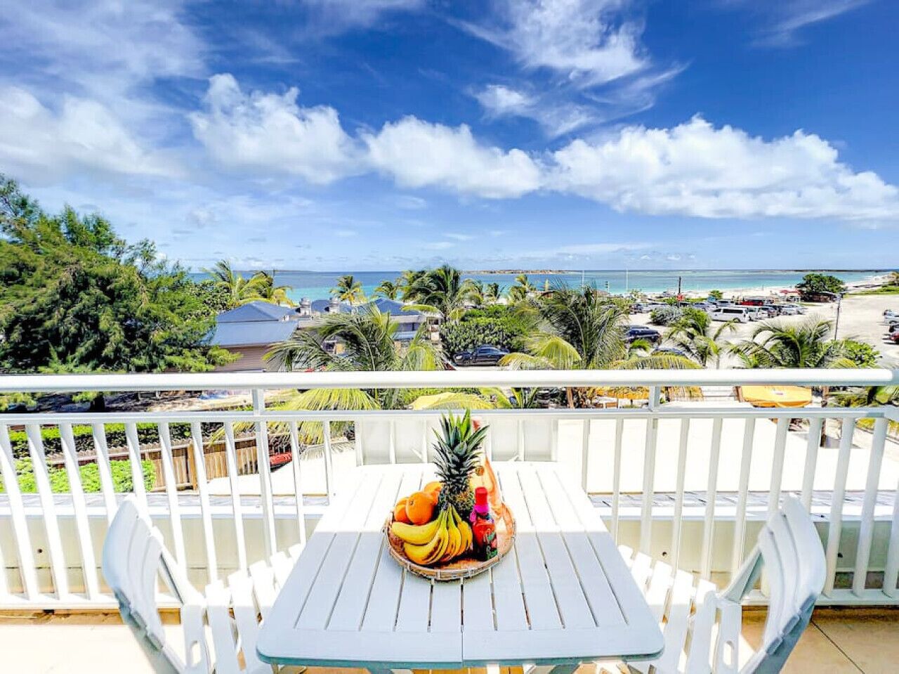A balcony with a table set for a tropical fruit drink, overlooking a beach and a parking area.