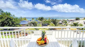 A balcony with a table set for a tropical fruit drink, overlooking a beach and a parking area.