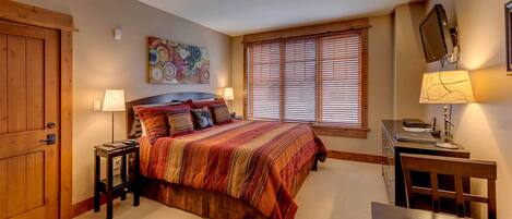 Een slaapkamer met een groot bed, twee nachtkastjes, een bureau met stoel, een televisie aan de muur, en een raam met zonwering.