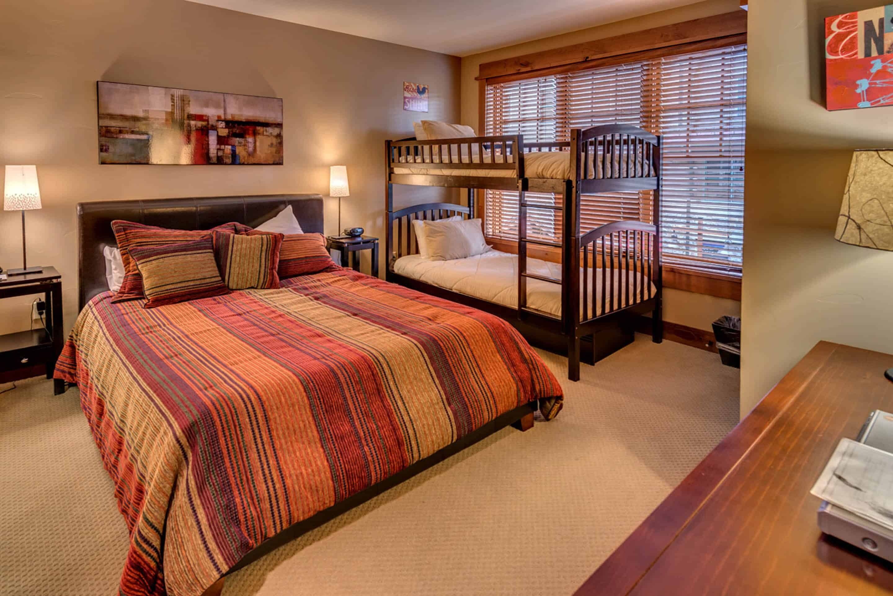 A bedroom with a large bed, a bunk bed, a desk, and a window with blinds.