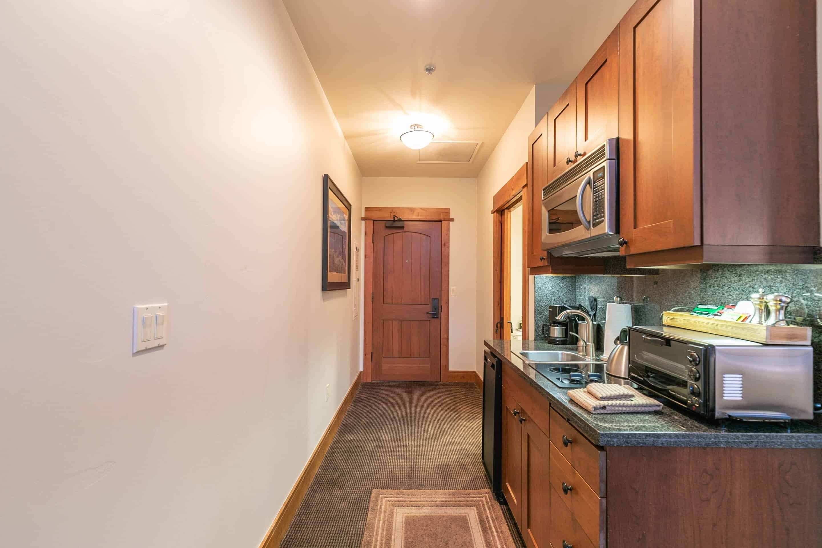 A narrow hallway leading to a wooden door, with a kitchen area featuring a microwave, toaster, and countertop with various items.